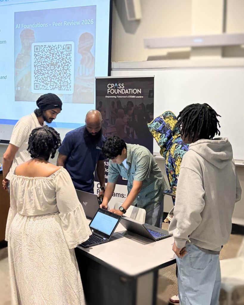 Group of participants gathered around a table in a classroom, working on laptops and a tablet. A projected slide reads “AI Foundations – Peer Review 2026” with a large QR code. A standing banner for “CPASS Foundation – Empowering Tomorrow’s STEMM Leaders” is visible behind the group. The setting includes a whiteboard, projector, and modern classroom equipment.