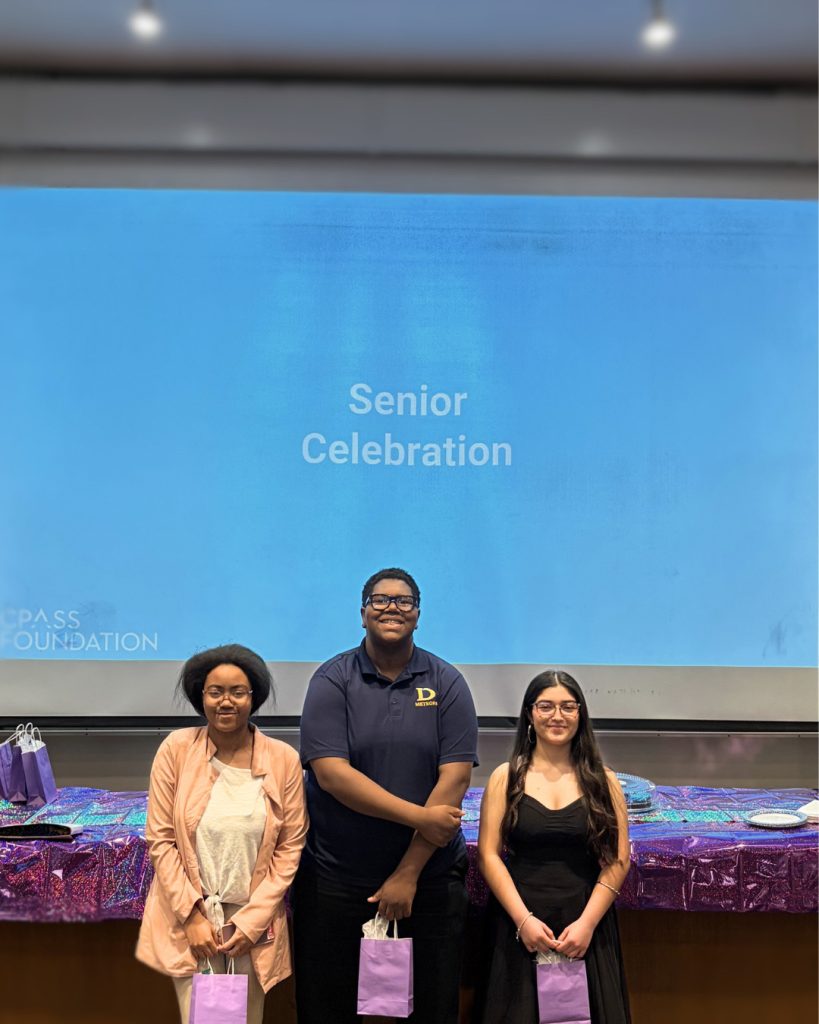 Three students stand at the front of a lecture hall holding small gift bags in front of a large projected slide that reads “Senior Celebration.” Behind them is a long table covered with a purple, reflective tablecloth, with plates, gift bags, and materials arranged across it. The setting includes a classroom with a projection screen, overhead lights, and presentation equipment, with a faint “CPASS Foundation” logo visible on the slide.
