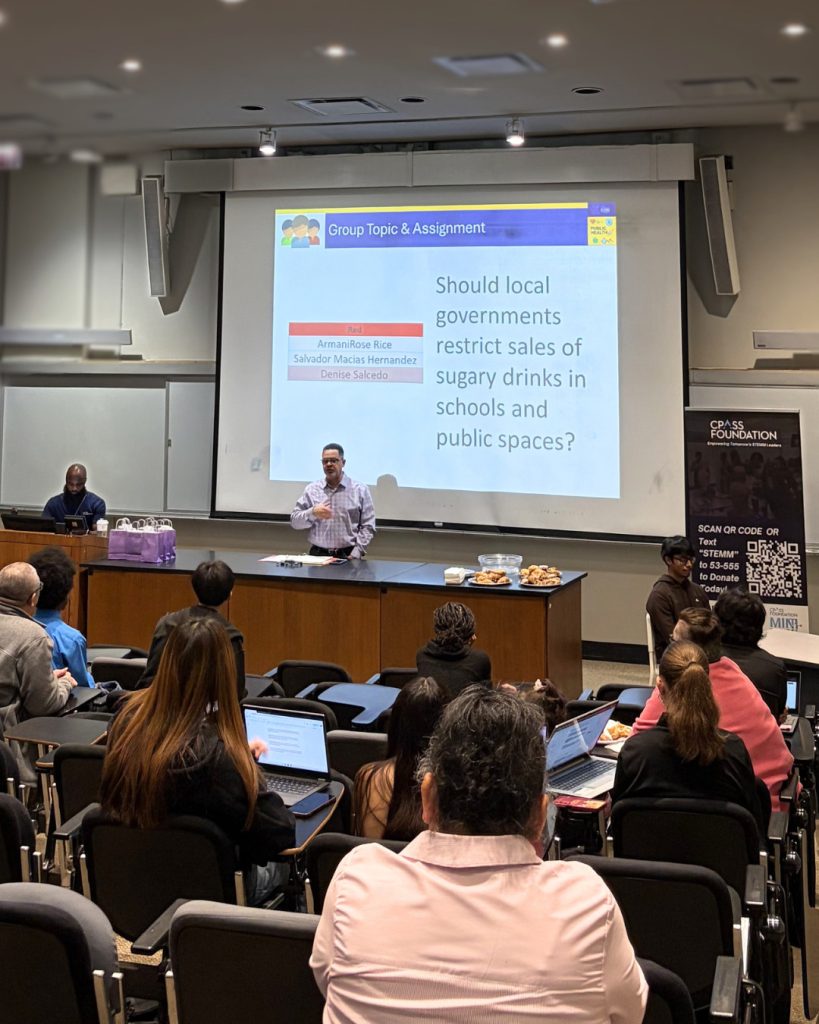 Lecture-style classroom with attendees seated and using laptops while a presenter stands at the front. A projected slide titled “Group Topic & Assignment” asks, “Should local governments restrict sales of sugary drinks in schools and public spaces?” A CPASS Foundation banner with a QR code stands to the side. A table at the front holds papers and trays of pastries.